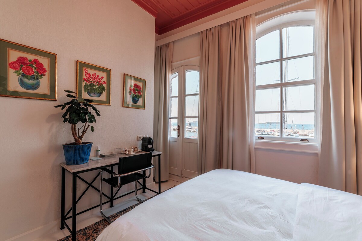 Ein Hotelzimmer mit Bett, Schreibtisch mit Stuhl, Blumentopf und Meerblick durch große Fenster.