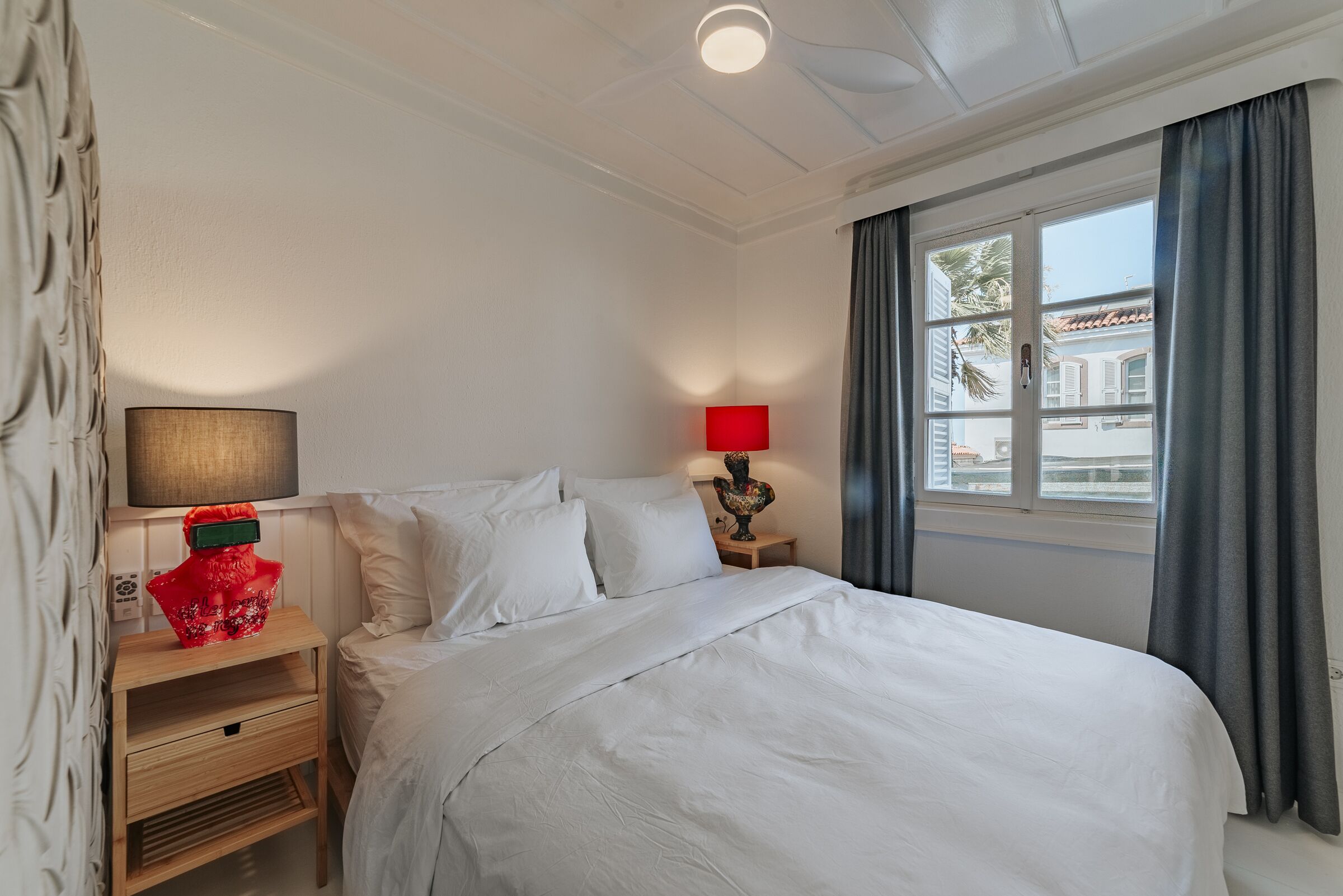 A neatly made bed with white linens, a wooden nightstand with a lamp, and a window with curtains.
