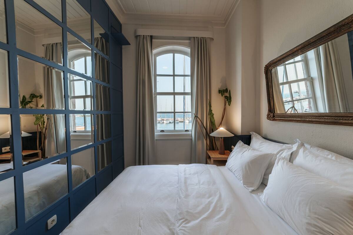 A hotel room with a large bed, a window with curtains, a mirror, and a view of the city.