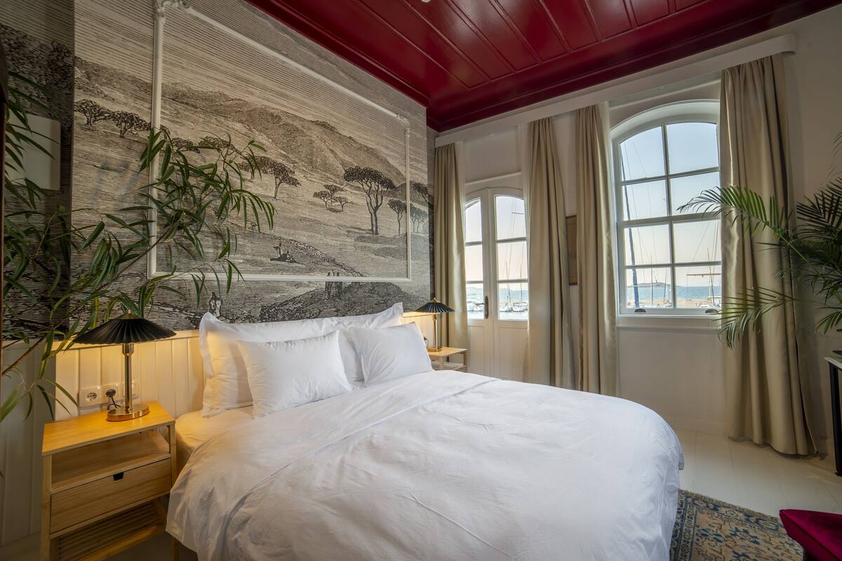 A neatly made bed with white linens, a nightstand with a lamp, a wall mural, and a large window with curtains.