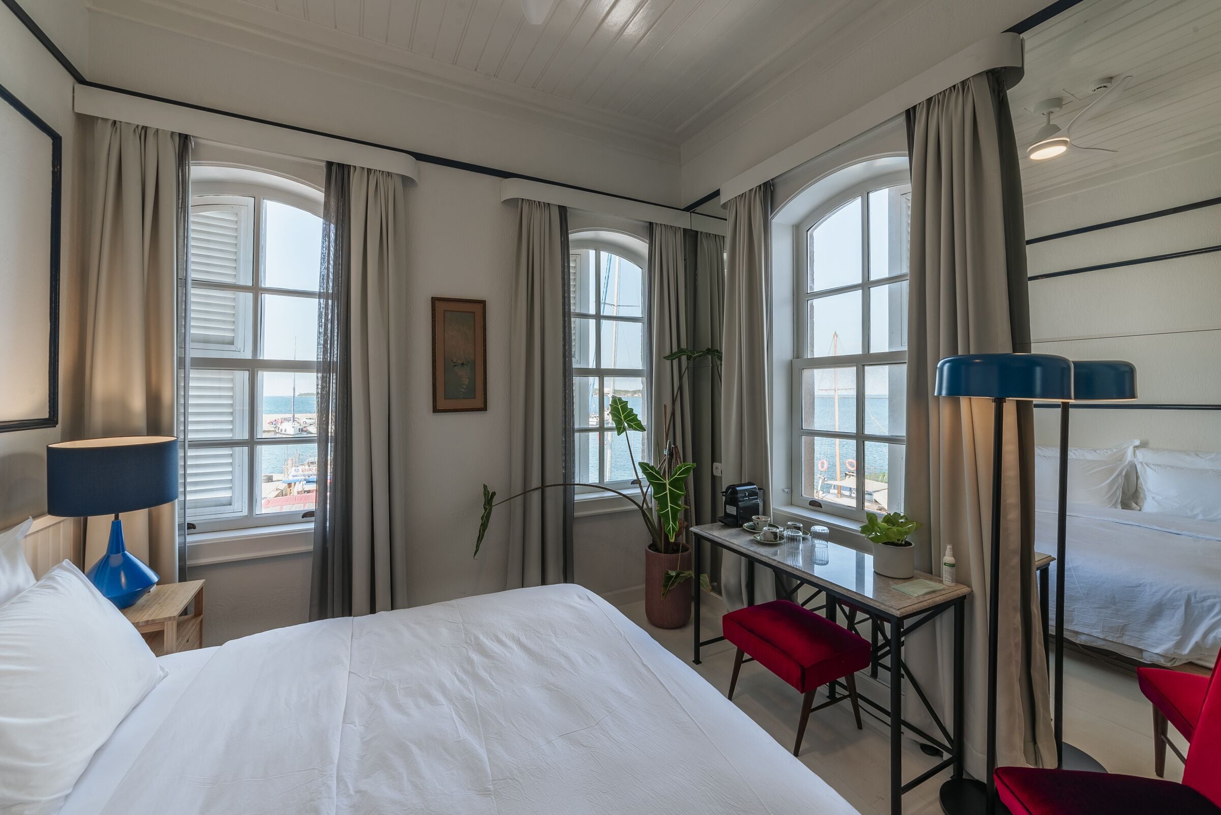 A hotel room with a bed, a desk, a chair, a lamp, a potted plant, and large windows with curtains.