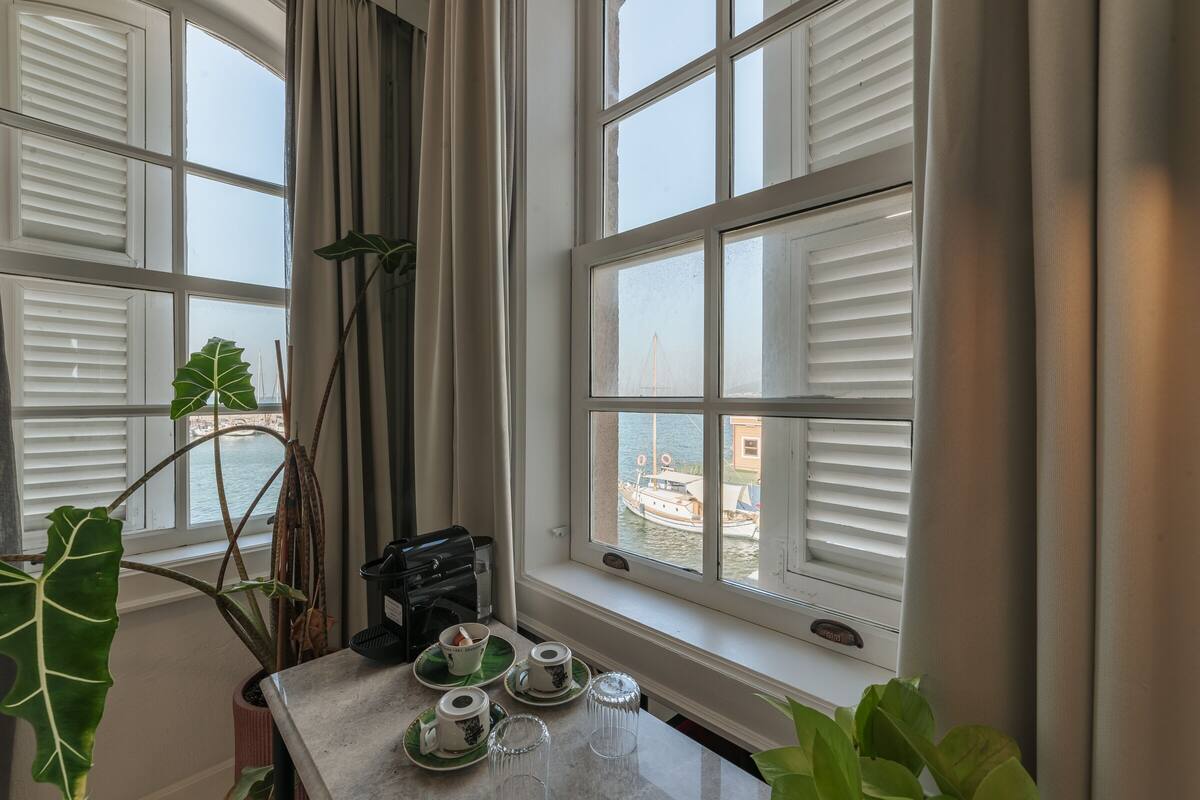 A room with a view of a body of water, a window with white shutters, a table with tea sets, and a potted plant.