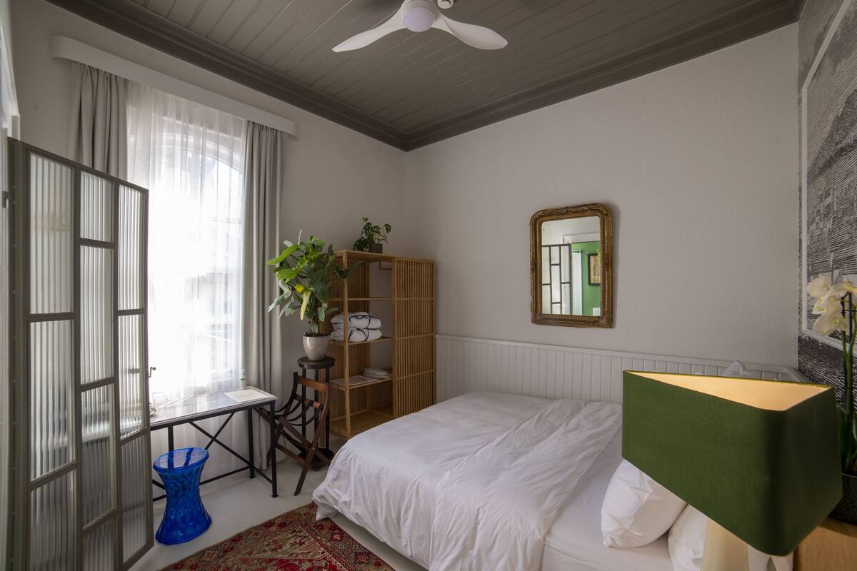A bedroom with a bed, a wooden wardrobe, a mirror, a window with curtains, a blue vase, and a potted plant.