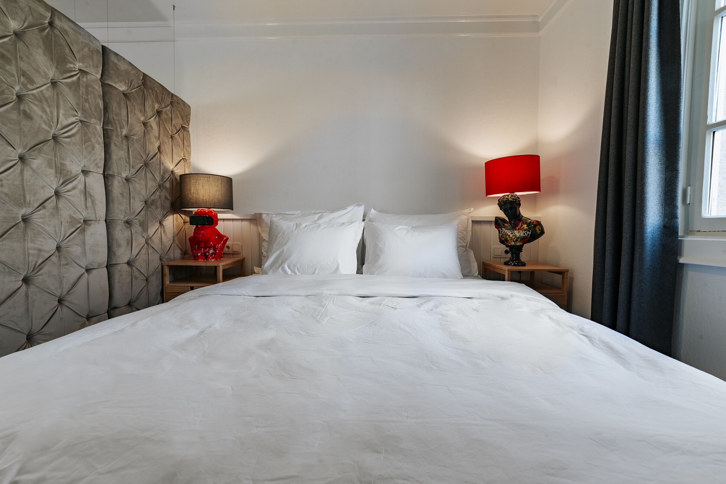 A hotel room with a tufted headboard, a large bed with white linens, two bedside tables with lamps, and a window with curtains.