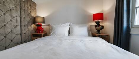 A hotel room with a tufted headboard, a large bed with white linens, two bedside tables with lamps, and a window with curtains.