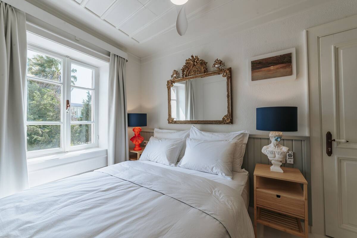 A neatly made bed with white linens, a large ornate mirror above it, and two bedside tables with lamps.