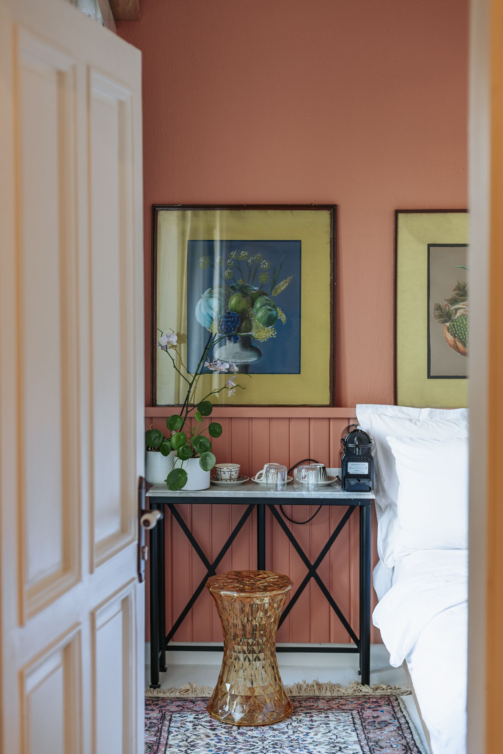 Una habitación con cama, una mesita con una planta, un cuadro enmarco y un taburete decorativo.