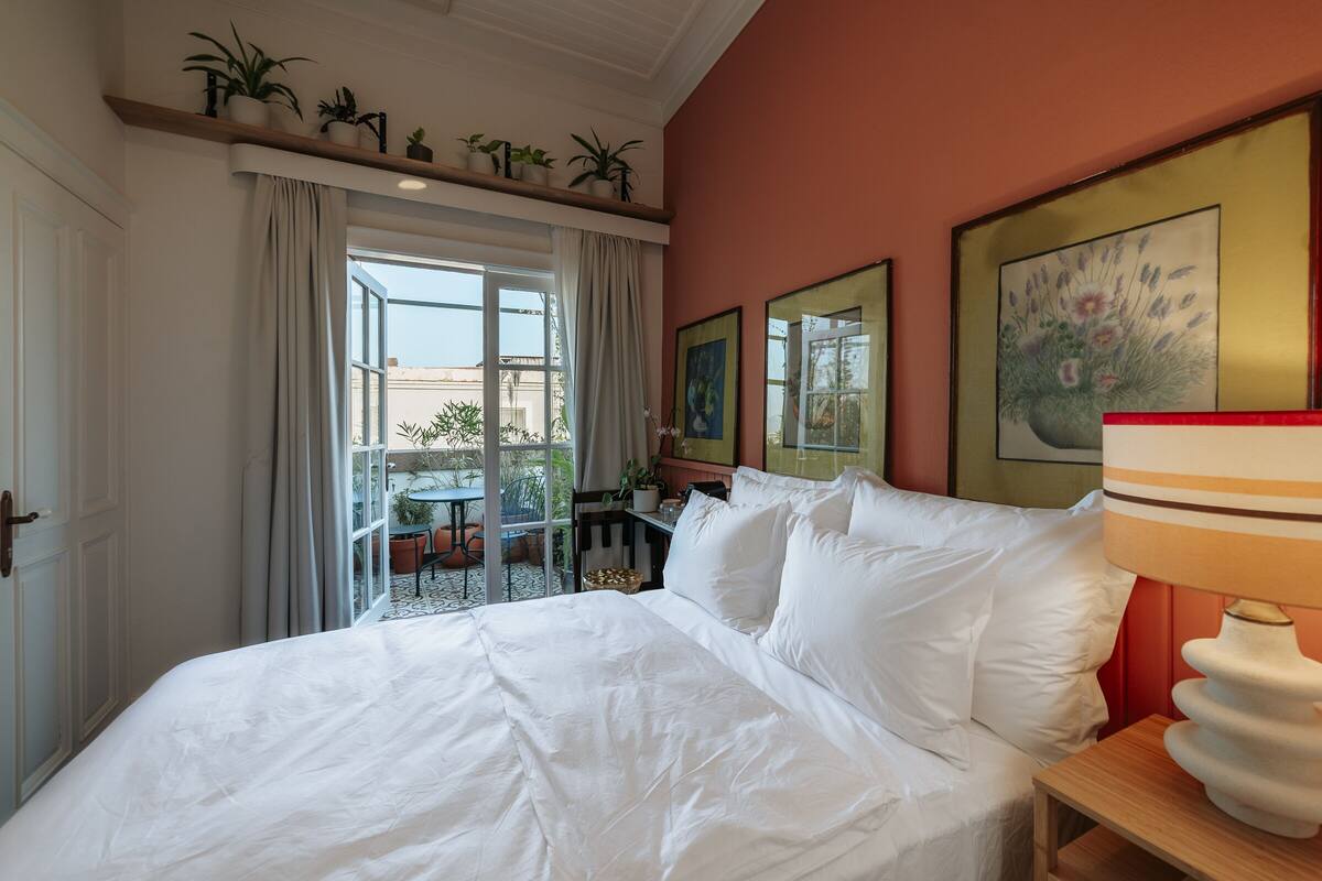 A hotel room with a large bed, a small table with a lamp, and a balcony with plants and a table.