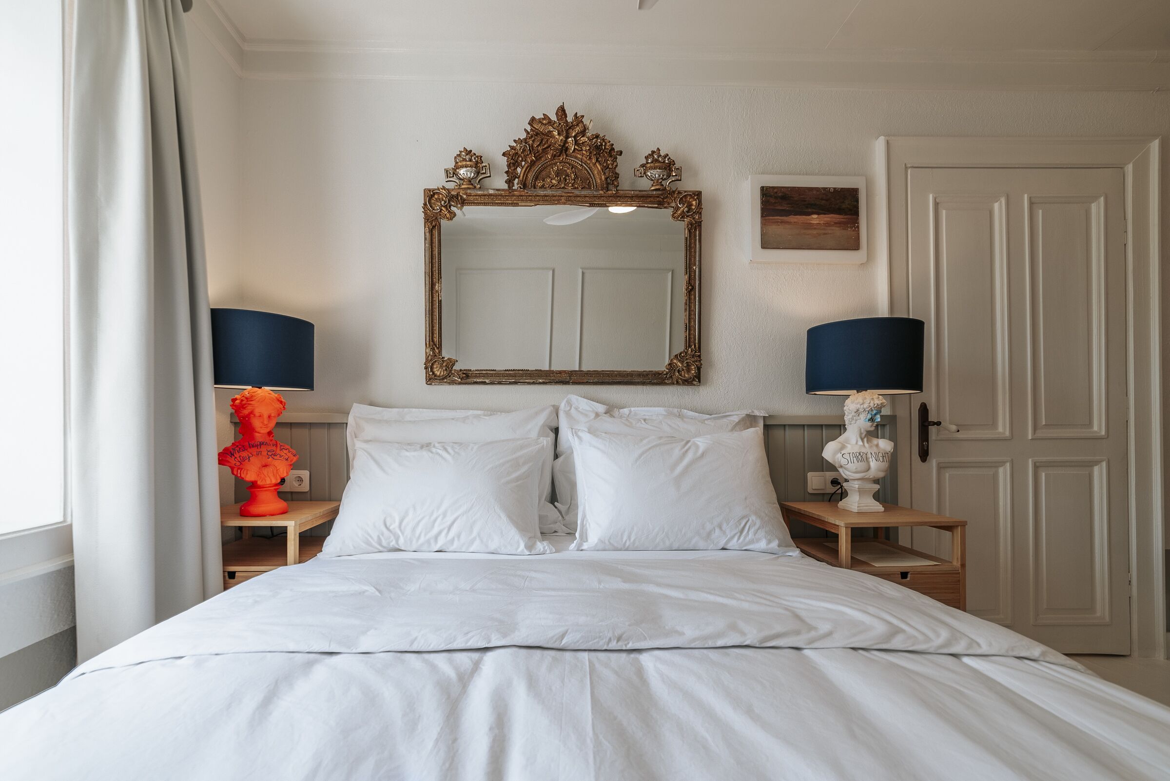 A neatly made bed with white linens, a large ornate mirror above it, and two bedside lamps on wooden nightstands.
