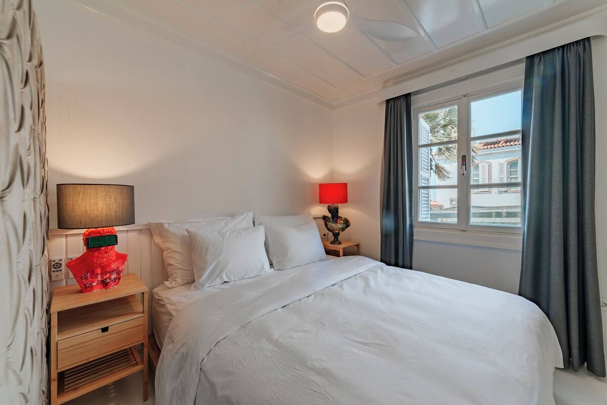 A neatly made bed with white linens, a wooden nightstand with a lamp, and a window with curtains.