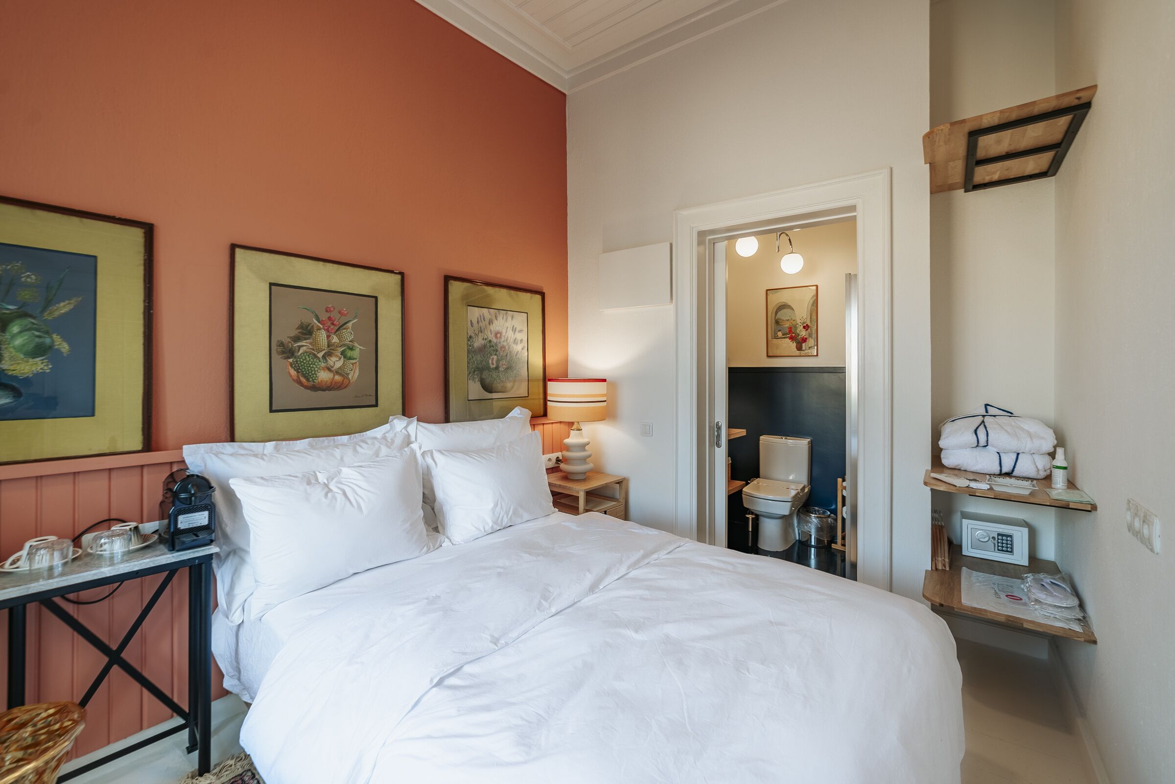 A hotel room with a large bed, a bedside table with a lamp, a small shelf with toiletries, and framed artwork on the wall.