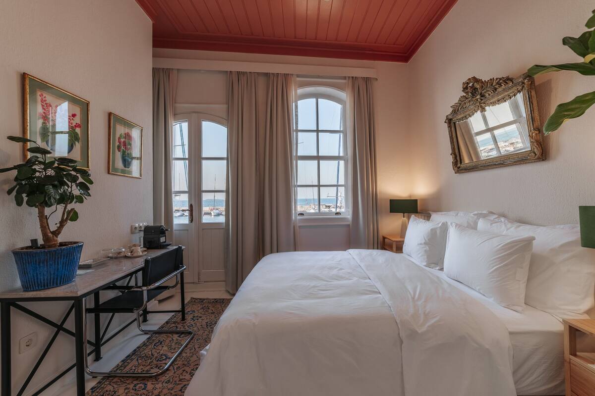 A hotel room with a bed, a desk with a chair, a potted plant, a window with curtains, and a view of the sea.