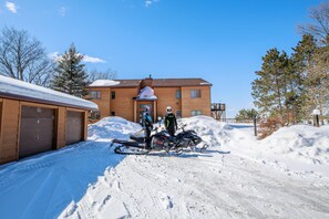 Une allée recouverte de neige, avec deux motoneiges garées devant un bâtiment doté d’un garage.