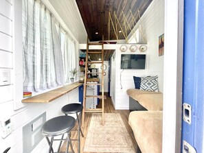 Interior compacto y bien organizado con techo de madera, una litera, un pequeño escritorio con taburetes y un televisor montado en la pared.