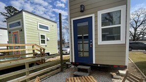 A small, single-story building with a blue door and windows, a wooden deck with a bench, and a gravel ground. - Tiny Home! With Great Layout!  Unique Stay Nurses Welcomed (Houston)