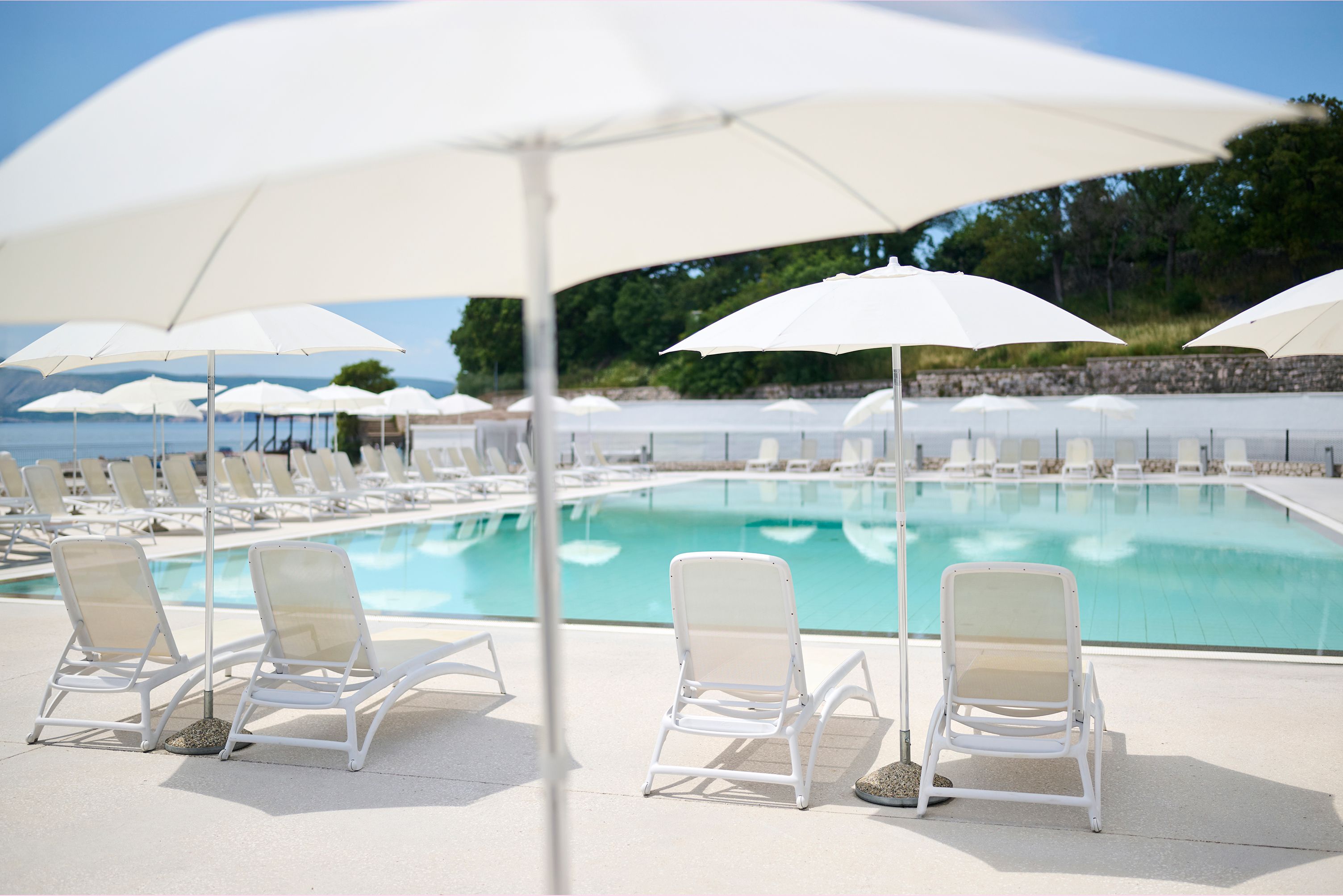 Un espace aménagé au bord de la piscine, avec des chaises longues blanches et des parasols, une piscine à l’eau claire et un mur de pierre en arrière-plan.