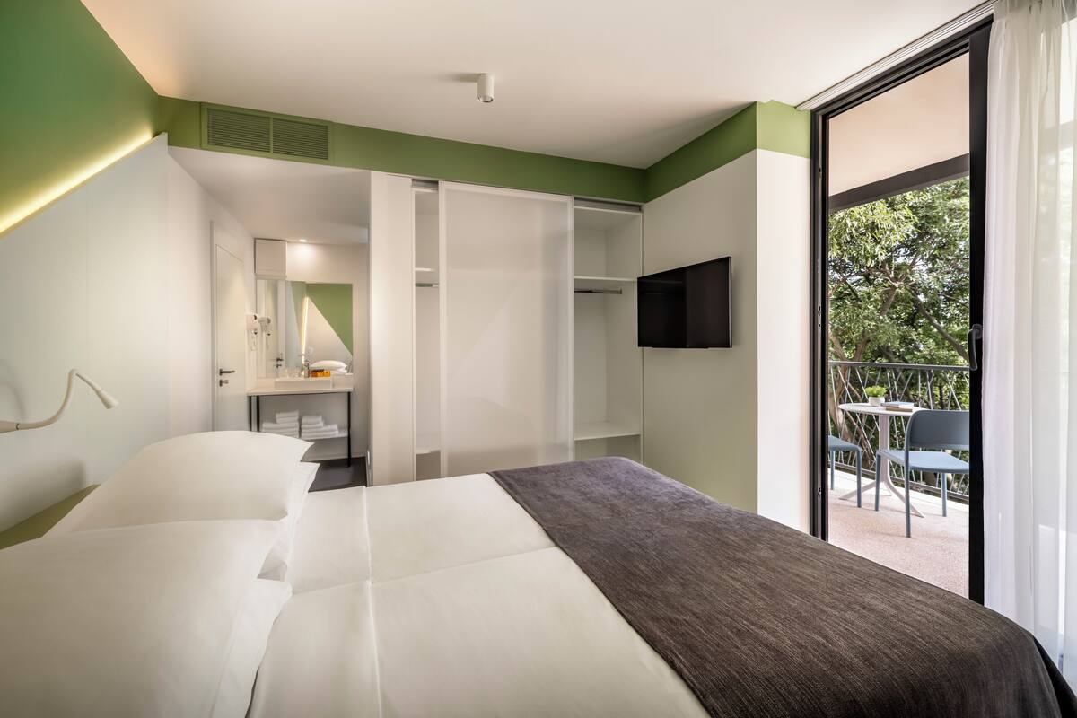 A hotel room with a bed, a bathroom with a mirror, a wardrobe, and a balcony with chairs.