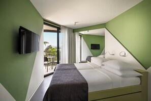 A modern hotel room with a large bed, a TV mounted on the wall, and a balcony with outdoor seating.