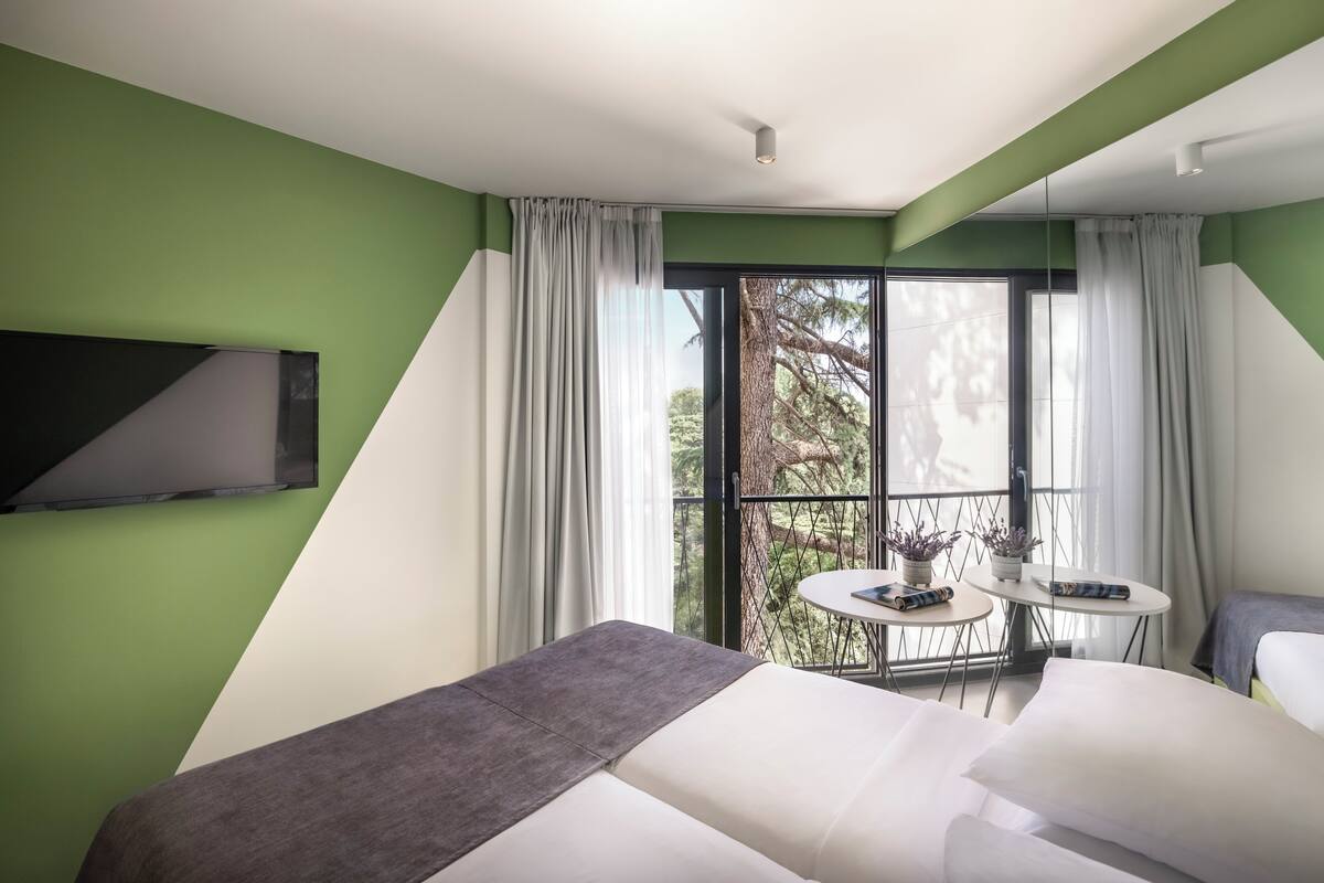 A modern hotel room with a large bed, a small table with a plant, and a balcony with a view of trees.