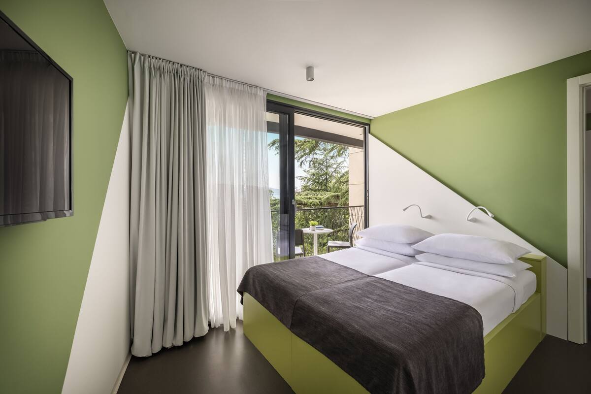 A modern bedroom with a bed, curtains, and a view of greenery outside.