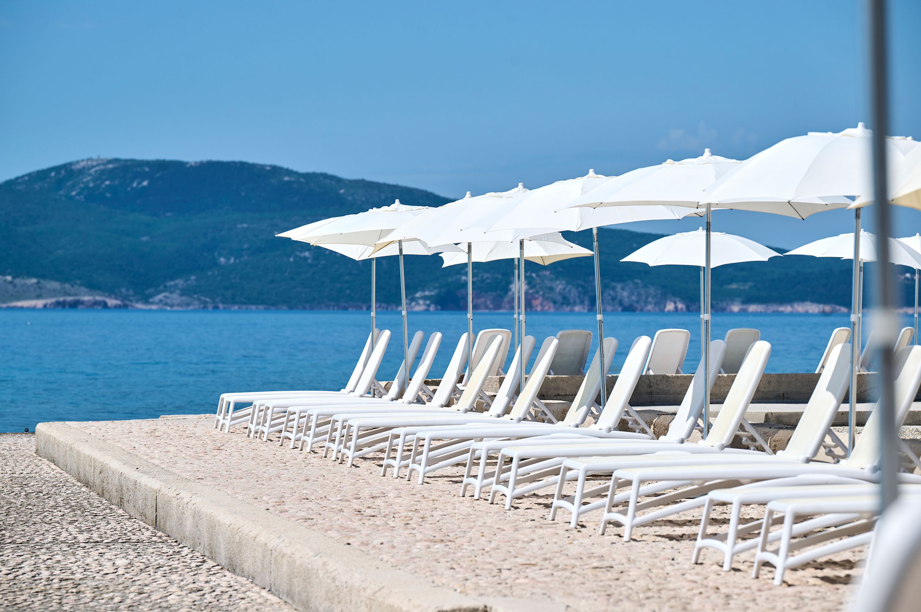 Une plage avec des chaises longues et des parasols blancs, une promenade en béton et une vue sur la mer et les montagnes.