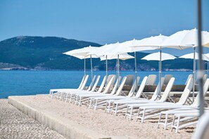 Une plage avec des chaises longues et des parasols blancs, une promenade en béton et une vue sur la mer et les montagnes.