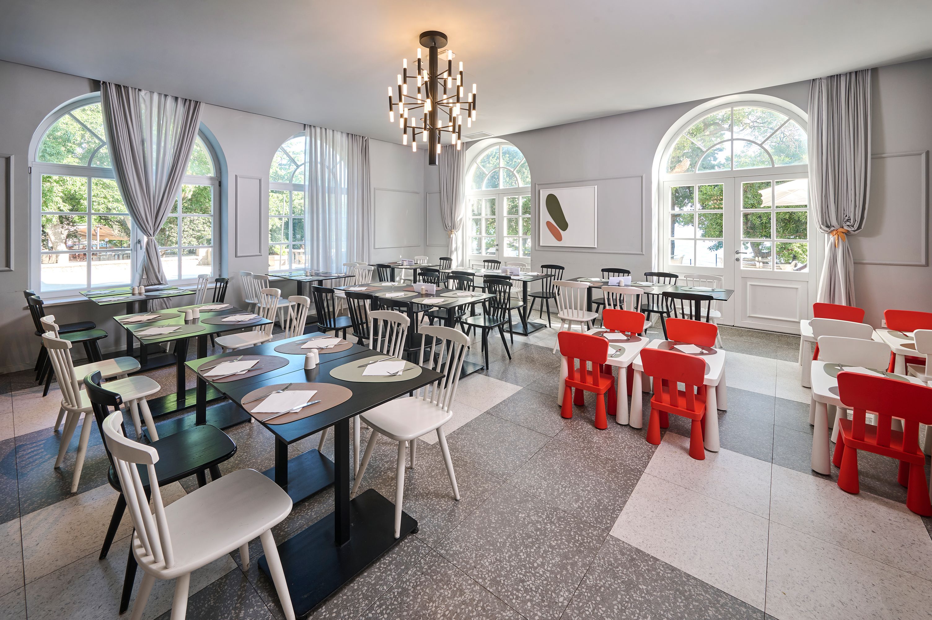 Una habitación amplia con varias mesas y sillas, ventanas grandes con cortinas y un candelabro.