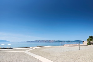 A beach volleyball court with a net, a pebble beach, and a clear view of the sea and distant mountains.