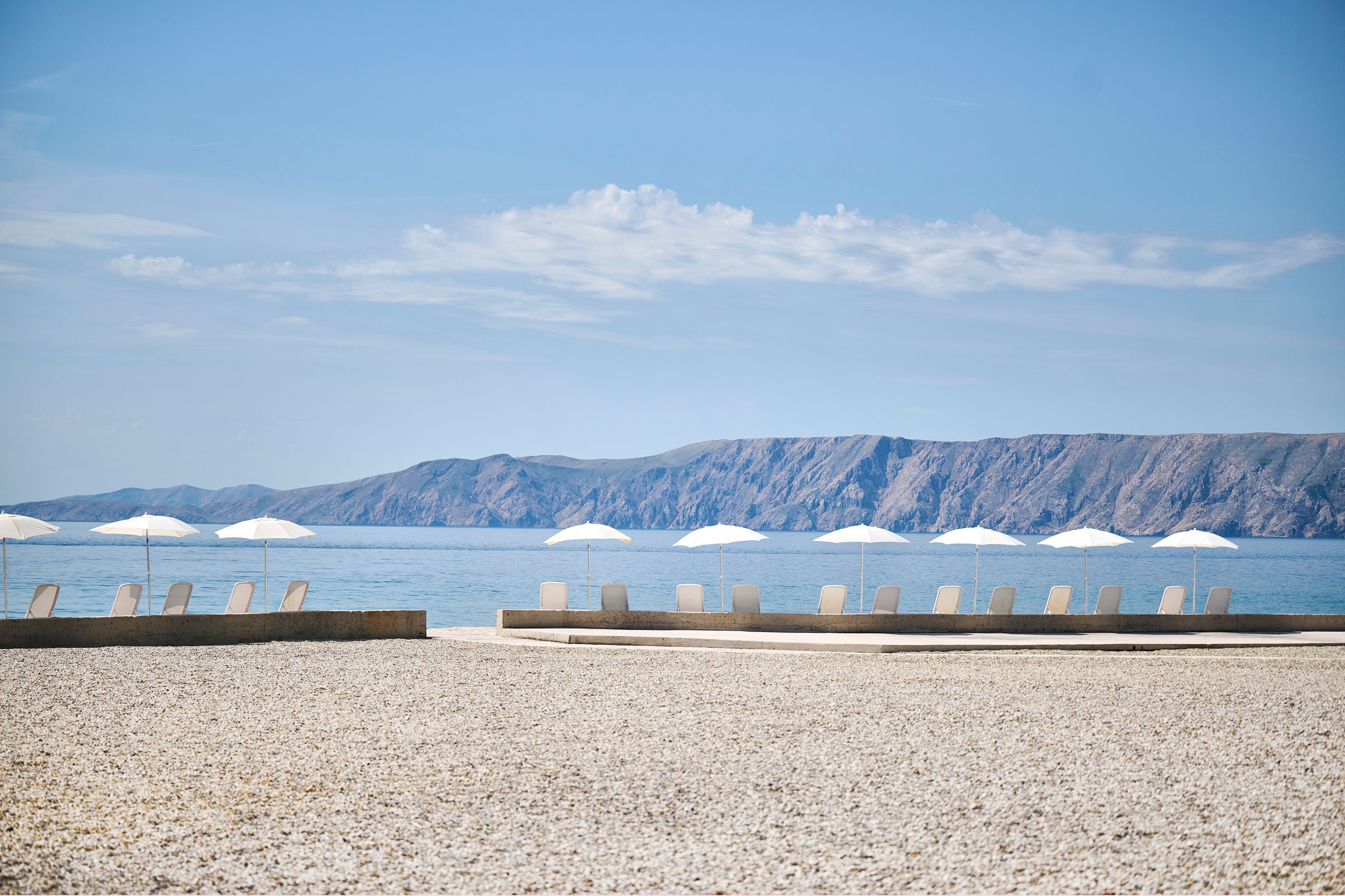 A pebble beach with lounge chairs and umbrellas, a calm sea, and mountains in the background.