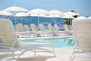 Un espace aménagé au bord de la piscine, avec des chaises longues blanches et des parasols, offrant une vue dégagée sur la mer et un paysage montagneux en toile de fond.