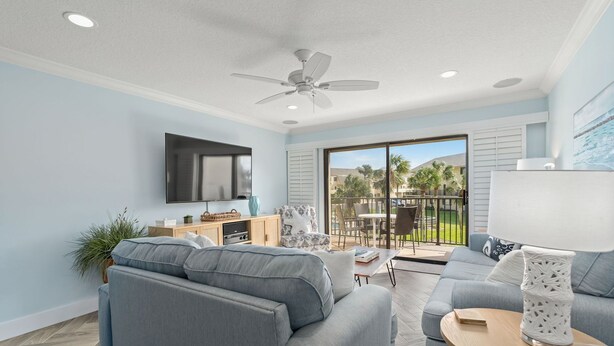 Ein modernes Wohnzimmer mit grauen Sofas, einem Flachbildfernseher, einem Deckenventilator und einem Balkon mit Sitzmöbeln.