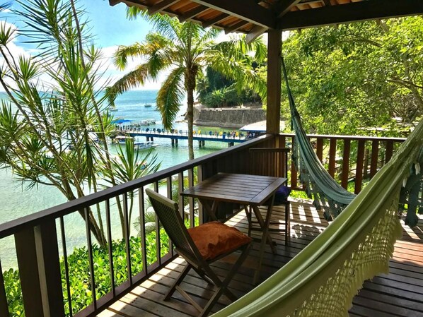 Une terrasse en bois avec un hamac, une table et une chaise, donnant sur une étendue d’eau où se trouvent des bateaux et, au loin, une île.
