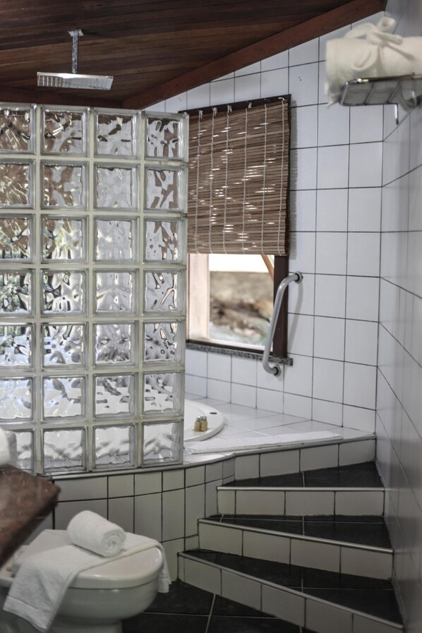 A bathroom with a toilet, a shower area, and a window with blinds. - Hotel Portaló (Cairu)