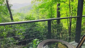 Un balcón con una silla de mimbre y una mesa de vidrio, con vistas a un bosque verde exuberante y colinas distantes.