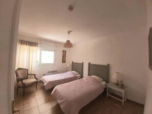 A bedroom with two beds, a chair, a bedside table, and a window with curtains.