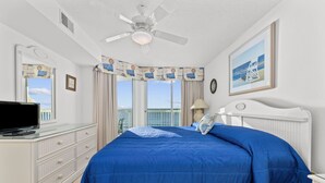 Un dormitorio con una cama grande, un ventilador de techo, un tocador, un televisor y una ventana con cortinas.