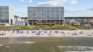 Un hotel frente a la playa con varios pisos, una playa de arena con sombrillas y vistas al océano.