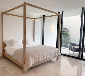 A four-poster bed with white bedding in a modern room with large windows and a view of the sea.