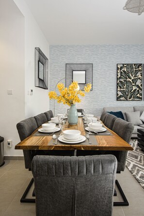 A dining area with a rectangular wooden table set for six, surrounded by grey upholstered chairs. A vase with yellow flowers is placed on the table.