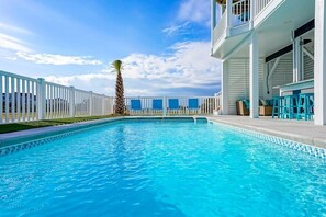 A swimming pool with a white fence, a palm tree, and a building with blue chairs and a balcony.