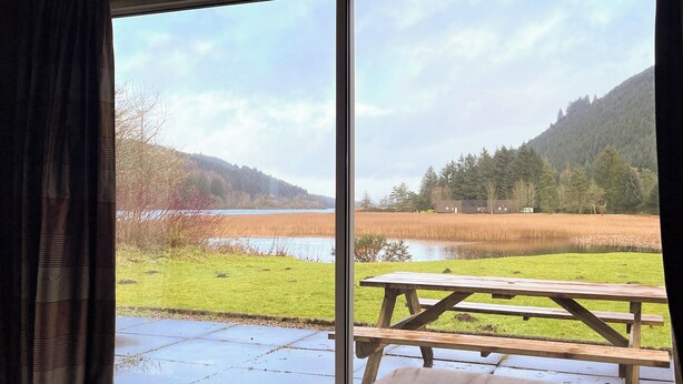 A view from a room with a large window overlooking a lake, picnic table, and forested hills.