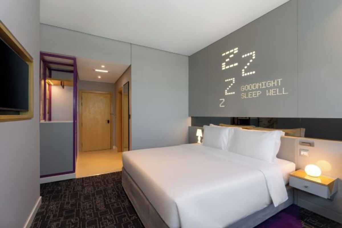 A modern hotel room with a large bed, a nightstand with a lamp, and a wall with illuminated text.