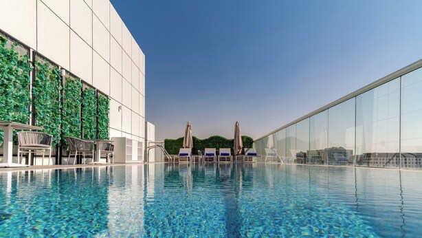 A modern indoor swimming pool with clear blue water, lounge chairs, and umbrellas, surrounded by glass walls and a building facade with greenery.