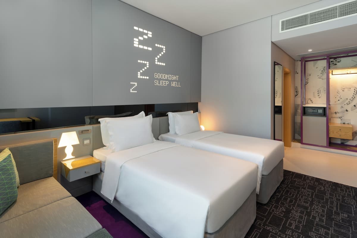 Una habitación de hotel moderna con dos camas individuales, una mesita de noche con lámpara, un pequeño espacio de descanso y un reloj de pared marcando las 22:00.