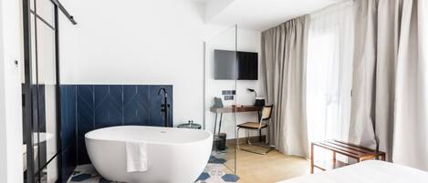 Una habitación de hotel moderna con una bañera grande, un escritorio con silla, una cama y un ventanal con cortinas.