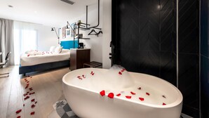 A modern hotel room with a large bathtub filled with water and rose petals, a bed with white linens, and a black tiled wall.