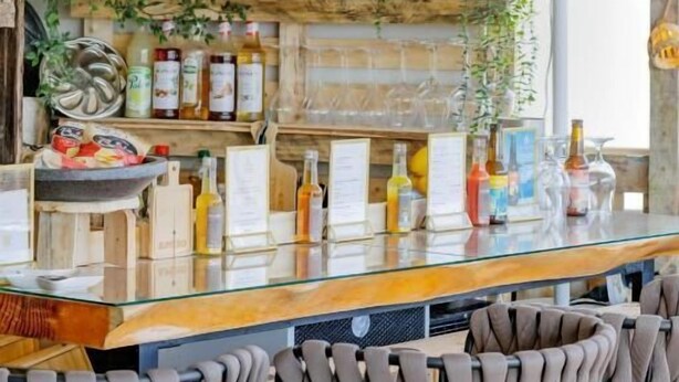 A bar area with wooden shelves stocked with bottles, a glass bar counter, and woven chairs.