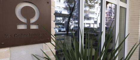 A sign with a symbol and text, a building entrance, and a plant in the foreground.