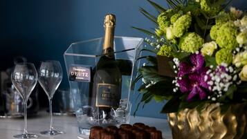 A champagne bottle, two glasses, and a plate of chocolates on a table with a decorative arrangement of flowers.