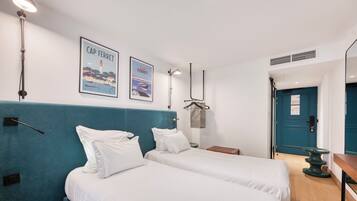 A hotel room with two beds, a blue headboard, white bedding, a wooden side table, a hanging coat rack, and two framed pictures on the wall.